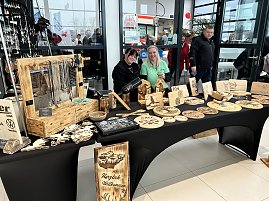 Zwei Frauen hinter einem Marktstand mit Holzgravuren und Schmuck, im Hintergrund Besucher im Autohaus.