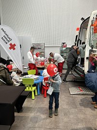 Kinder basteln an einem Tisch mit Rotkreuz-Ballons, daneben Sanit&auml;ter und ein offener Rettungswagen im Autohaus.