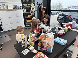 Eine Frau macht einen Sehtest mit einem kleinen M&auml;dchen. Daneben sitzt ein kleiner Junge. Auf dem Tisch davor liegen Flyer, eine Box und Material.