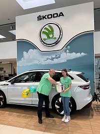 Mann und junge Frau in gr&uuml;nen Shirts stehen vor einem wei&szlig;en �koda im Autohaus. Die junge Frau h&auml;lt einen silbernen Beh&auml;lter, der Mann greift hinein und zieht einen Umschlag.