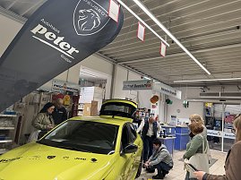 Gelber Peugeot im Baumarkt, eine Frau wechselt ein Rad, daneben mehrere Zuschauer.
