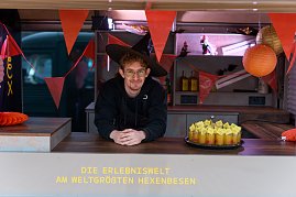 Junger Mann im Foodtruck mit Hexenhut, Halloween-Deko und Tablett mit Shots.