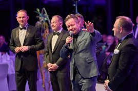 Redner mit Mikrofon vor elegant gekleideten Gästen bei Gala-Event.