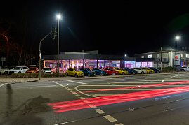 Außenaufnahme bei Nacht: Ein modern verglastes Autohaus ist festlich beleuchtet, innen findet eine Veranstaltung statt. Davor stehen mehrere Fahrzeuge in einer Reihe.