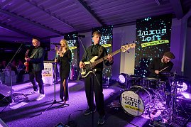 Eine Live-Band sorgt für Stimmung: Sängerin, Gitarrist, Bassist und Schlagzeuger stehen auf einer Bühne mit moderner Lichttechnik und performen vor den Gästen. Im Hintergrund mehrere Displays mit der Aufschrift Luxus Loft Die Party WG.