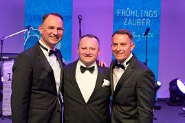 Drei Herren im Smoking stehen beieinander vor der Bühne und schauen lächelnd in die Kamera.