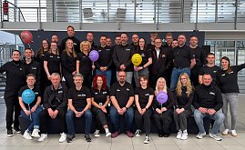 Man sieht ein Teamfoto mit vielen Menschen auf und vor einer kleinen B&uuml;hne. Alle haben das gleiche T-Shirt an, manche halten bunte Luftballons hoch.