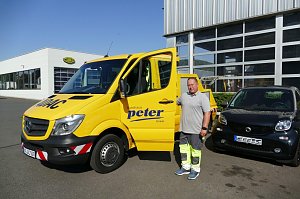 Ein Mitarbeiter des Autohaus Peter steht neben einem gelben ADAC-Servicefahrzeug mit geöffneter Tür. Im Hintergrund eine moderne Werkstatthalle mit Glasfronten und ein schwarzes Smart-Auto, links ein weiteres Gebäude.