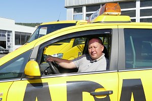 Ein Mitarbeiter des Autohaus Peter sitzt am Steuer eines gelben ADAC-Servicefahrzeugs mit großem ADAC-Schriftzug. Er schaut aus dem offenen Fenster und lächelt, im Hintergrund sind weitere gelbe Servicefahrzeuge sowie eine Werkstatthalle mit großen Fenstern zu sehen.