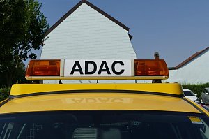 Nahaufnahme eines gelben Fahrzeugs mit ADAC-Schriftzug auf dem Dachbalken mit orangefarbenen Warnleuchten. Im Hintergrund sind Häuser mit hellen Fassaden sowie Bäume zu sehen.