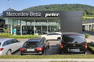 Mercedes-Benz Autohaus Peter mit Glasfassade und gro&szlig;em Logo, davor geparkte Mercedes-Fahrzeuge, im Hintergrund gr&uuml;ne bewaldete H&uuml;gel und blauer Himmel.