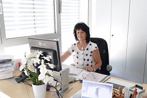 Frau mit dunklen Haaren sitzt an einem Schreibtisch in einem hellen B&uuml;ro. Sie arbeitet am Computer und schaut in die Kamera. Auf dem Schreibtisch stehen eine Orchidee, Akten und B&uuml;romaterial.