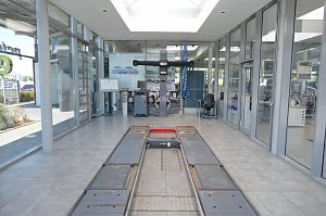 Helles, modernes Mercedes-Benz-Autohauses mit großen Glasfronten. In der Mitte eine Fahrzeughebebühne im Boden eingelassen, dahinter Werkstattausrüstung und ein Schreibtisch mit Stuhl. Rechts Glasbüros mit Blick in den Innenraum.