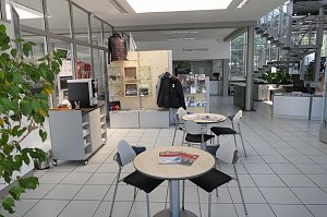 Heller, moderner Warte- und Servicebereich des Autohauses. Im Vordergrund runde Tische mit St&uuml;hlen, Zeitschriften darauf. Im Hintergrund eine Verkaufsfl&auml;che mit ausgestellten Jacken und Zubeh&ouml;r, eine Treppe f&uuml;hrt ins obere Stockwerk. Viel Glas und Licht, Pflanzen sorgen f&uuml;r eine freundliche Atmosph&auml;re.