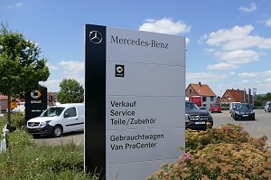 Gro&szlig;es Mercedes-Benz Schild auf einem Autohaus-Gel&auml;nde. Auf dem Schild stehen die Angebote: Verkauf, Service, Teile/Zubeh&ouml;r, Gebrauchtwagen, Van ProCenter. Im Hintergrund mehrere Mercedes-Fahrzeuge, ein weiteres Smart-Schild und rote Hausd&auml;cher. Blauer Himmel mit einigen Wolken.