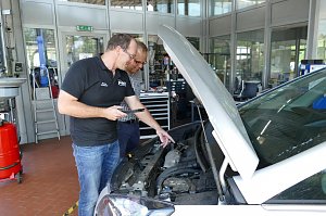Zwei Männer stehen in einer Werkstatt neben einem Auto mit geöffneter Motorhaube. Der Mann links trägt ein schwarzes Poloshirt mit "Peter" Logo und zeigt mit einem Tablet auf den Motor. Der andere Mann trägt ein kariertes Hemd und schaut interessiert. Im Hintergrund sieht man Werkstatt-Ausrüstung, Werkbänke und Glaswände mit Blick in weitere Bereiche der Werkstatt.
