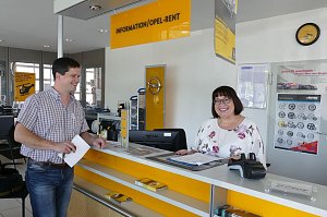 Ein Mann und eine Frau stehen am Empfangstresen mit der Aufschrift INFORMATION / OPEL-RENT. Die Frau hinter dem gelb-weißen Tresen trägt eine Brille und ein geblümtes Oberteil, sie lächelt. Der Mann vor dem Tresen hält Unterlagen in der Hand und lächelt ebenfalls. Im Hintergrund hängen Opel-Plakate, es ist ein gelbes Opel-Logo an der Wand sichtbar. Der Empfangsbereich wirkt hell und freundlich.