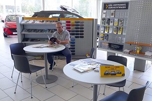 Ein Mann sitzt in einer hellen Opel-Ausstellungshalle an einem Tisch und liest eine Broschüre. Im Hintergrund sind Regale mit Prospekten, Farbmustern und Zubehör zu sehen. Rechts befindet sich eine Tafel mit dem Hinweis Opel zertifizierte Gebrauchtwagen. Auf den Tischen liegen Zeitschriften und Flyer. Es sind mehrere schwarze Stühle um die Tische angeordnet.