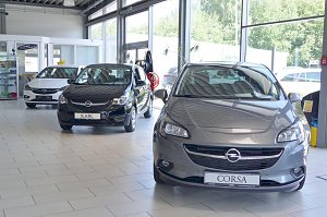 In einem hellen Autohaus-Ausstellungsraum stehen drei Opel-Fahrzeuge. Im Vordergrund ein grauer Opel Corsa, daneben ein schwarzer Opel Karl und im Hintergrund ein weißes Fahrzeug. Der Raum hat große Fenster, durch die viel Tageslicht einfällt. Im Hintergrund sind Glaskabinen und Vitrinen mit Zubehör zu sehen.