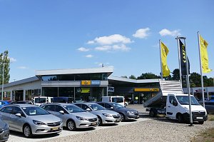 Außenansicht des Opel-Autohauses in Osterode mit großem Vorplatz. Im Vordergrund stehen mehrere silberfarbene Opel-Pkw und ein weißer Opel-Transporter mit Kipper-Aufbau. Vor dem Gebäude wehen gelbe und schwarze Opel-Flaggen. Das Autohaus hat eine moderne Glasfassade, darüber sind Schilder mit Opel Service und dem Opel-Logo sichtbar. Der Himmel ist blau mit einigen kleinen Wolken.