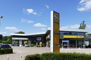 Außenansicht eines Opel-Autohauses in Osterode. Das moderne Gebäude hat eine große Glasfront und ein markantes, gelb-blaues Opel-Logo auf einem hohen Pylon im Vordergrund. Vor dem Autohaus sind mehrere Fahrzeuge geparkt. Der Himmel ist blau mit einigen kleinen Wolken, wodurch die Szene freundlich und einladend wirkt.