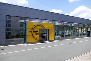 Eingang des Opel-Autohauses Peter in Sondershausen. Die Glasfassade des modernen Geb&auml;udes spiegelt die Umgebung wider, w&auml;hrend der gelbe Opel-Eingangsbereich mit gro&szlig;em Markenlogo und Willkommen-Schriftzug hervorsticht. Durch die Fenster sind Autos im Innenraum sichtbar.