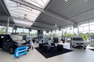 Großer, heller Showroom eines Mercedes-Benz Autohauses mit hohen Glasfronten und modernen Deckenleuchten. Mehrere Vans und Transporter sind ausgestellt, darunter ein weißer und ein schwarzer Mercedes. In der Mitte ein schwarzer Teppich mit vier schwarzen Sesseln und einem Tisch als Wartebereich. Viel natürliches Licht durch die großen Fenster.