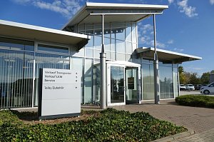 Glasfassade des Mercedes-Autohauses in Zerbst mit modernem Eingangsbereich, daneben ein Wegweiser mit Hinweisen zu Verkauf, Service und Zubehör, davor niedrige Bodendecker-Pflanzen, blauer Himmel im Hintergrund.