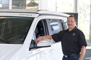 Ein Autohaus-Mitarbeiter im schwarzen Hemd mit weißem Mercedes-Logo steht lächelnd neben einem weißen Westfalia-Campervan, lehnt mit einer Hand an der Fahrertür.