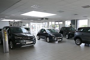 Heller, moderner Showroom des Opel-Autohauses mit mehreren ausgestellten Autos. Links vorne steht ein grauer Opel Mokka X, daneben ein kleinerer Opel Adam in der Mitte. Im Hintergrund sind weitere Fahrzeuge und Fahrräder zu sehen. Die großen Fensterfronten lassen viel Tageslicht herein, die Decke ist mit weißen Paneelen und Einbaulampen ausgestattet. An der Rückwand hängen Farbmuster.