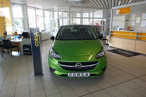Heller Verkaufsraum des Opel-Autohauses mit einem grünen Opel Corsa im Vordergrund. Links stehen Schreibtische mit Stühlen und Informationsmaterialien. Im Hintergrund sieht man den Empfangsbereich, an der eine Mitarbeiterin sitzt. Große Fenster lassen viel Tageslicht herein, und die Decke ist mit weißen Paneelen versehen. Ein Schild mit der Aufschrift "CORSA" steht neben dem Auto.