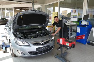Ein Mechaniker überprüft einen silbernen Opel mit offener Motorhaube in einer Werkstatt. Der Mechaniker steht freundlich lächelnd neben dem Fahrzeug und bedient ein Prüfgerät für die Scheinwerfer. Im Hintergrund sind Schreibtische, Poster und technische Geräte sichtbar.