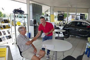 Zwei Männer in einem hellen Autohaus-Innenraum beim Handschlag. Der ältere Mann sitzt am Tisch, trägt ein kariertes Hemd und Shorts, der jüngere Mann in rotem Polo-Shirt steht und hält Dokumente. Im Hintergrund sind ein schwarzes Auto, Tische, Prospektständer und Regale mit Zubehör und Modellautos zu sehen. Große Fenster lassen viel Tageslicht herein.