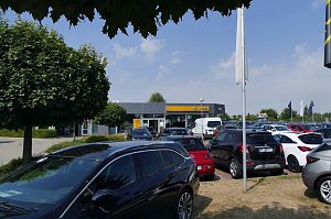 Blick über den Parkplatz eines Opel-Autohauses in Sömmerda. Im Vordergrund stehen viele verschiedene Fahrzeuge, darunter schwarze, rote und weiße Modelle. Im Hintergrund ist das moderne Autohaus mit der markanten gelben Opel-Beschilderung zu sehen, umgeben von Bäumen und Flaggen. Der Himmel ist blau mit ein paar Wolken.