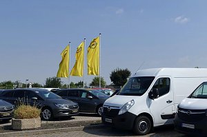 Auf dem Autohausgelände in Sömmerda stehen mehrere Opel-Fahrzeuge, darunter ein weißer Transporter und mehrere graue PKW. Im Hintergrund wehen drei große gelbe Fahnen mit dem Opel-Logo. Der Himmel ist blau mit einigen leichten Wolken.
