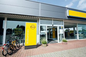 Eingang des Opel-Autohauses in Sömmerda. Links stehen zwei Fahrräder an einem Geländer. Im Vordergrund ein gelbes Schild mit dem Opel-Logo und der Aufschrift Willkommen. Der Eingang ist verglast mit automatischen Türen, daneben stehen Pflanzkübel. Das Gebäude wirkt modern, mit großen Glasfronten und einem grauen Dach.