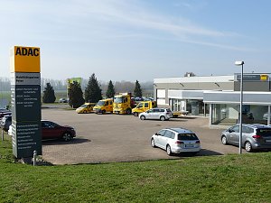 Alt-Text: Blick &uuml;ber das Gel&auml;nde des ADAC Mobilit&auml;tspartners Peter in Nordhausen. Im Vordergrund ein gro&szlig;es gelbes ADAC-Schild mit Serviceleistungen, dahinter mehrere gelbe ADAC-Fahrzeuge und silberne Mietwagen, im Hintergrund das moderne Geb&auml;udekomplex mit Glasfassaden und Werkstatt.