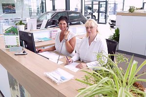 Zwei Mitarbeiterinnen an der Rezeption des Škoda-Autohauses, eine telefoniert, die andere schreibt. Helle, moderne Umgebung mit Pflanzen und Autos im Hintergrund.