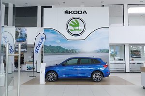 Innenansicht des Škoda-Autohauses mit großem weißen Škoda-Logo an der Wand. In der Mitte steht ein blauer Škoda Scala, flankiert von Werbefahnen und Postern.