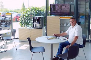 Ein &auml;lterer Mann sitzt l&auml;chelnd an einem runden Tisch in einer hellen Lounge im Opel-Autohaus Peter in Nordhausen. Auf dem Tisch liegen Zeitschriften. Im Hintergrund stehen ein Kaffeeautomat, ein TV mit Nachrichten und Prospektst&auml;nder. Durch die gro&szlig;en Fenster sieht man Autos und gr&uuml;ne Pflanzen drau&szlig;en.