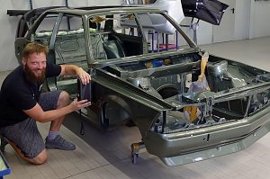 Tino Schwenzer kniet l&auml;chelnd neben einer teilweise montierten gr&uuml;nen Autokarosserie in einer Werkstatt. Er h&auml;lt ein Messger&auml;t an die Seitenwand, um den Farbton zu bestimmen. Im Hintergrund verschiedene Autoteile und Werkstattausr&uuml;stung. 