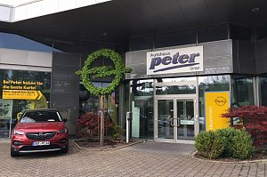 Eingang des Autohauses Peter GmbH, Osterode. Über dem Glasdoppeltür-Eingang ist ein großes Schild mit dem Schriftzug Autohaus Peter GmbH angebracht. Daneben steht ein roter Opel. Auffällig ist ein großes Opel-Logo aus grünem Buchsbaum. Rechts vom Eingang befindet sich ein gelbes Willkommen-Schild mit Opel-Logo.