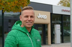 Ihr Verkaufsberater f&uuml;r die amerikanischen Fabrikate: Danny Kindervater mit gr&uuml;ner Jacke steht l&auml;chelnd vor dem Jeep-Autohaus, im Hintergrund das Eingangsportal mit Jeep-Logo.