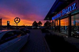Mercedes-Benz Autohaus bei Sonnenuntergang. Der Himmel leuchtet in kräftigen Orange- und Lilatönen. Auf dem Parkplatz stehen mehrere Autos, der beleuchtete Mercedes-Stern und der Schriftzug ‚Mercedes-Benz‘ an der Glasfassade reflektieren das Licht.