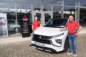 Zwei Mitarbeiter in roten Mitsubishi-Poloshirts stehen vor einem wei&szlig;en Mitsubishi Eclipse Cross Plug-in Hybrid, vor dem Eingang des Autohauses Peter in G&ouml;ttingen.