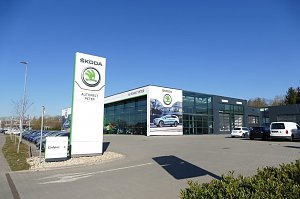 Modernes Škoda-Autohaus Autowelt Peter mit großem Glasfassaden-Design, hohem weißen Škoda-Schild und mehreren geparkten Autos bei blauem Himmel.