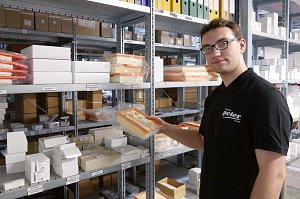  Ein junger Mann mit Brille und schwarzem Autohaus Peter-Poloshirt steht im Ersatzteillager. Er h&auml;lt einen in Plastik verpackten Luftfilter in der Hand. Im Hintergrund sind zahlreiche Regale mit Autoersatzteilen, Kartons und Ordnern zu sehen.