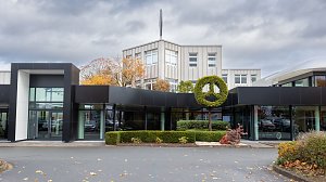 Frontansicht des modernen Mercedes-Autohauses in Nordhausen mit gro&szlig;en Glasfl&auml;chen, schwarzer Fassade und einem Mercedes-Benz Logo als Formschnitt-Hecke, herbstliche B&auml;ume im Hintergrund, bew&ouml;lkter Himmel.