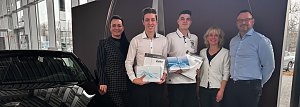 Christina Dost, Gesch&auml;ftsf&uuml;hrerin, steht links neben Paul W. (zweiter von links), der nach verk&uuml;rzter Ausbildung als Assistent der Gesch&auml;ftsleitung startet, und Domenik B. (Mitte), der das Team k&uuml;nftig als Karosseriebauer verst&auml;rkt. Rechts daneben stehen Silke Lang, Ausbildungsleiterin, und Enrico Dost, Serviceleiter. Paul W. und Domenik B. halten jeweils eine Geschenkbox mit hellblauer Schleife in den H&auml;nden. Die Gruppe steht im Autohaus vor einem schwarzen Auto und einer Wanddekoration und l&auml;chelt in die Kamera.