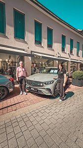 Zwei Personen posieren vor einem grauen Mercedes-Ausstellungswagen, geparkt vor einem Bekleidungsgesch&auml;ft.